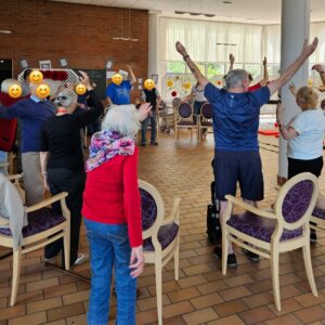 Group of seniors in a community room standing and raising arms in a seated/standing exercise class, with chairs around and large windows in the background.