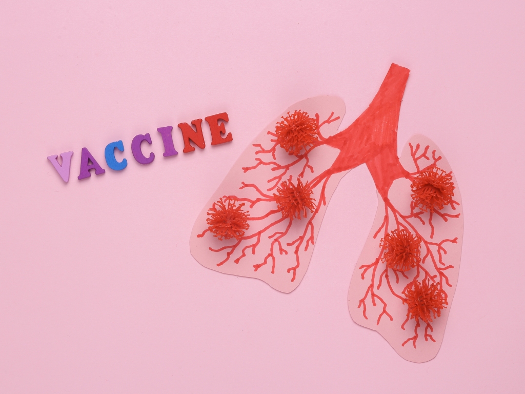 Pink background with the word VACCINE in colorful letters beside a cut-out lungs diagram showing red bronchial branches and virus-like particles.