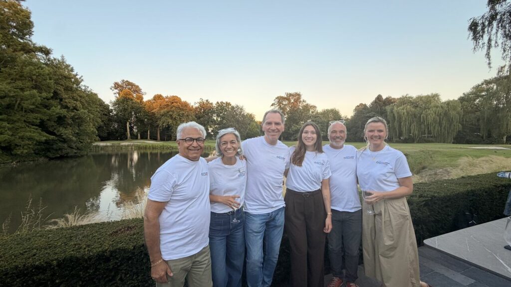Six people wearing white T-shirts pose together by a lake in a park at sunset, smiling for a group photo.