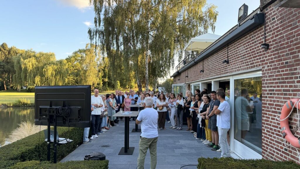 Large group of people gathered on a patio by a pond, listening to a man in a white shirt speak at a standing table outside a brick building.