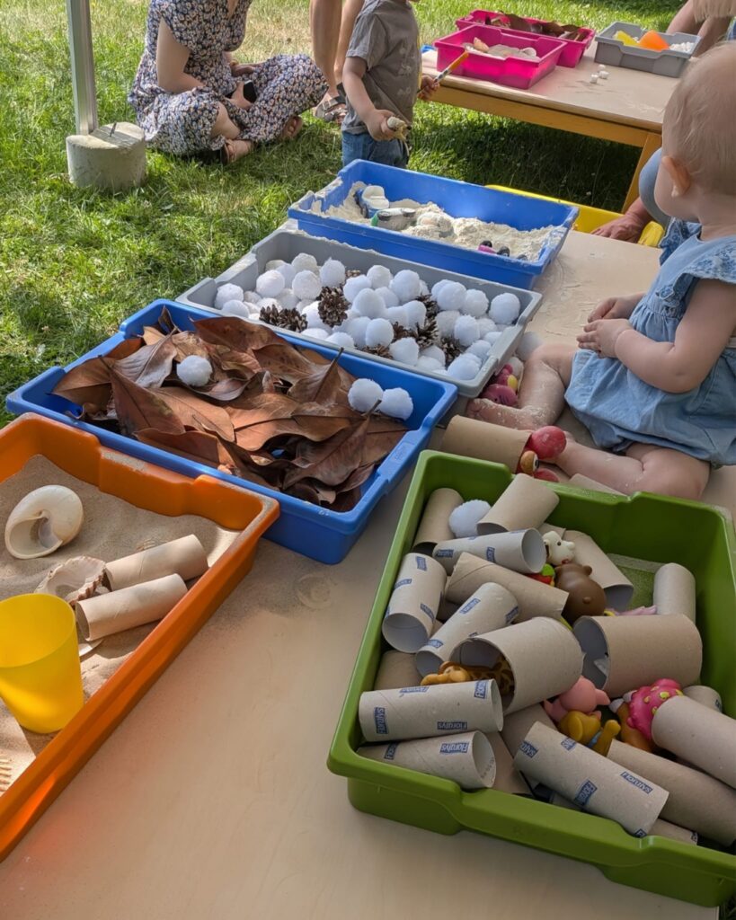 Children at a sunny outdoor play table with labeled sensory bins filled with leaves, pinecones, white balls, and cardboard tubes for digging and sorting.