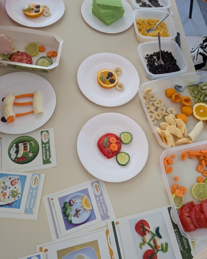 Top-down view of a children’s snack table with white plates of fruit artistry and trays of cornflakes, raisins, and sliced fruit like orange, kiwi, and banana.
