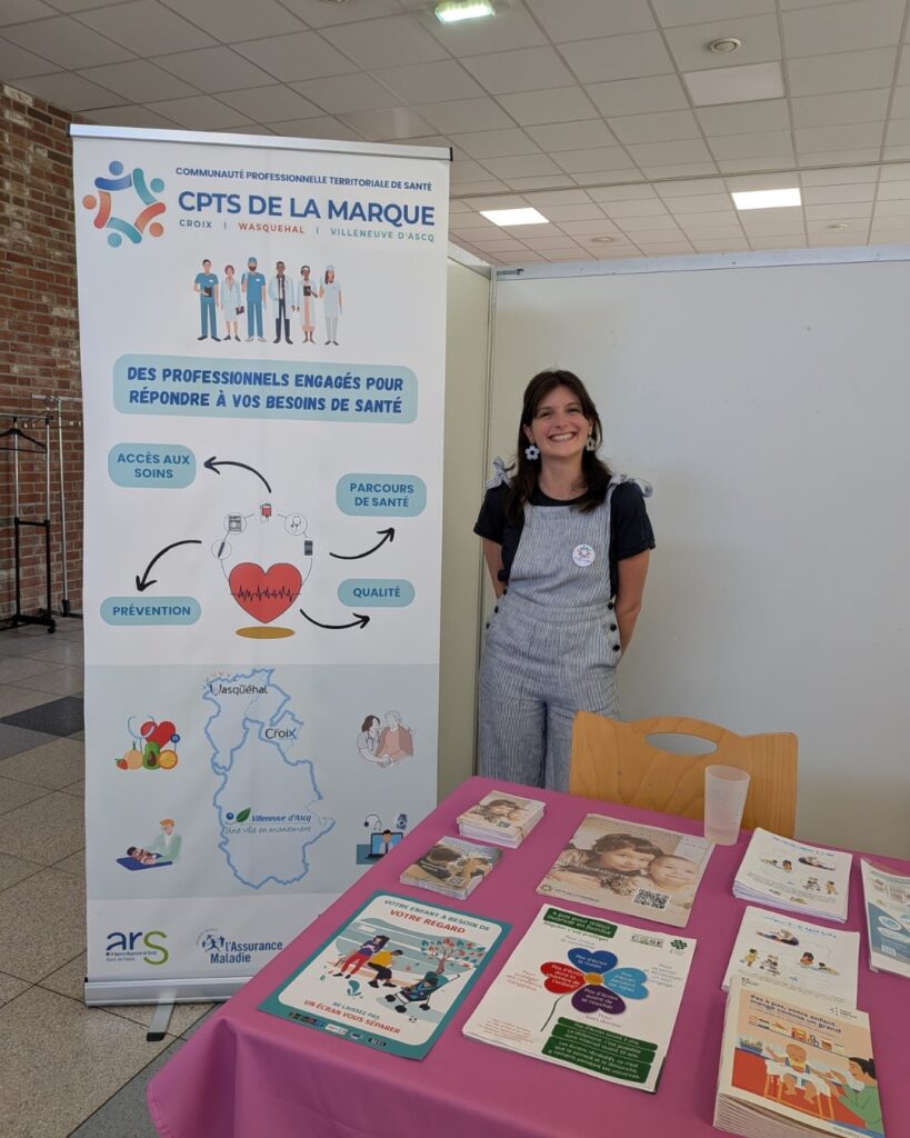 Smiling woman in striped overalls stands beside a health information banner at a booth.